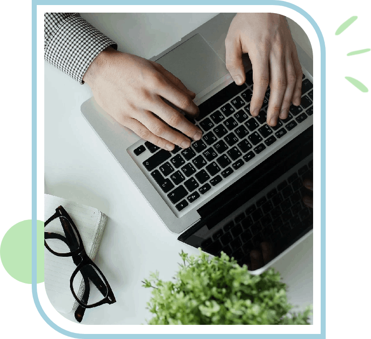 A person typing on a laptop with glasses and plant in the background.